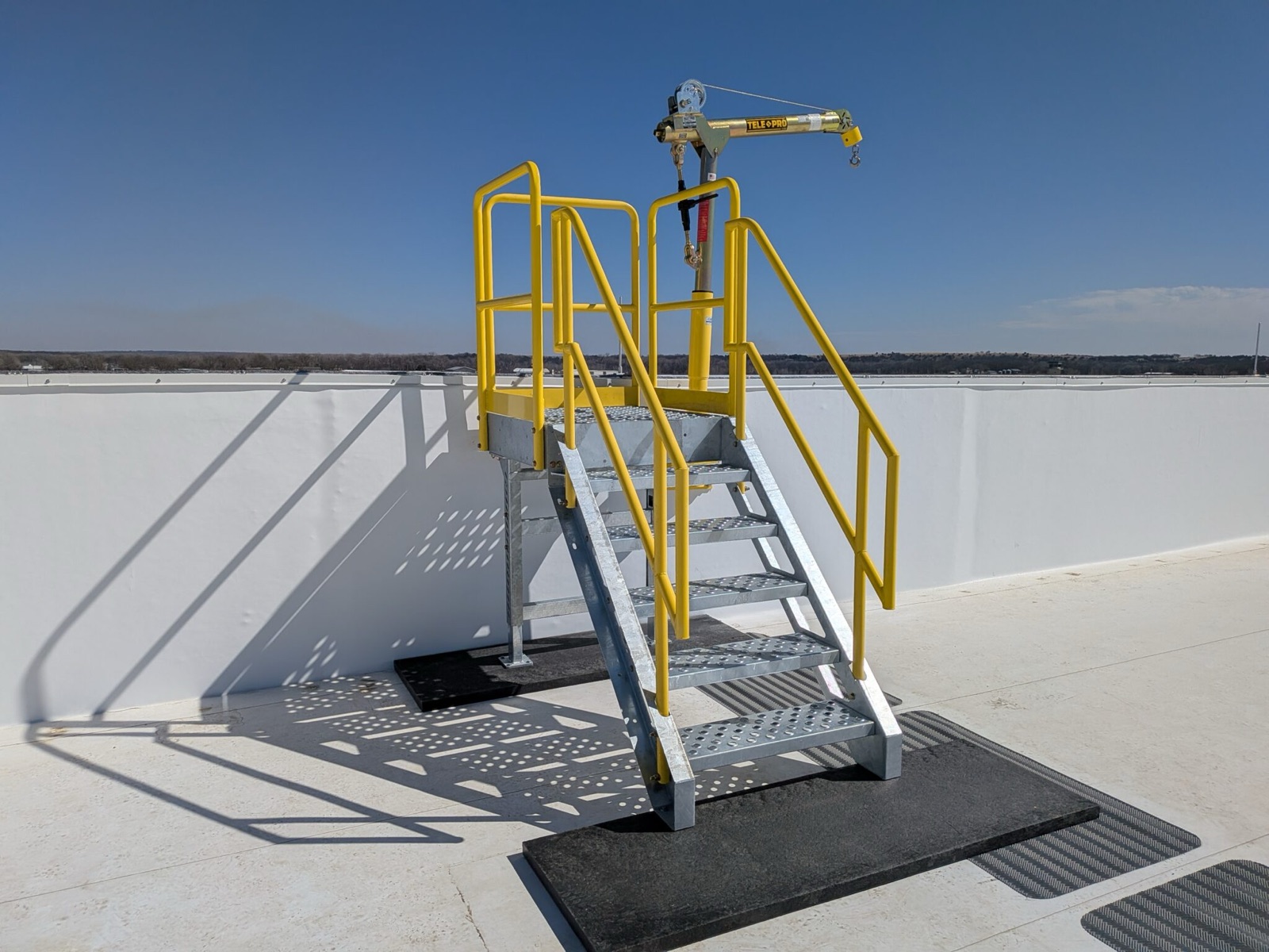 A modular crossover stair on non-penetrating padded feet over a roof parapet, with a davit material lift at the top